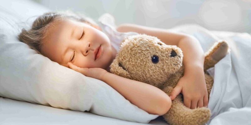 Mädchen schläft friedlich im Bett und kuschelt mit einem Teddybär: Symbolbild für guten Kinderschlaf vor dem Schulstart, entspannte Abendroutine und Schlafrituale für Kinder in Deutschland.